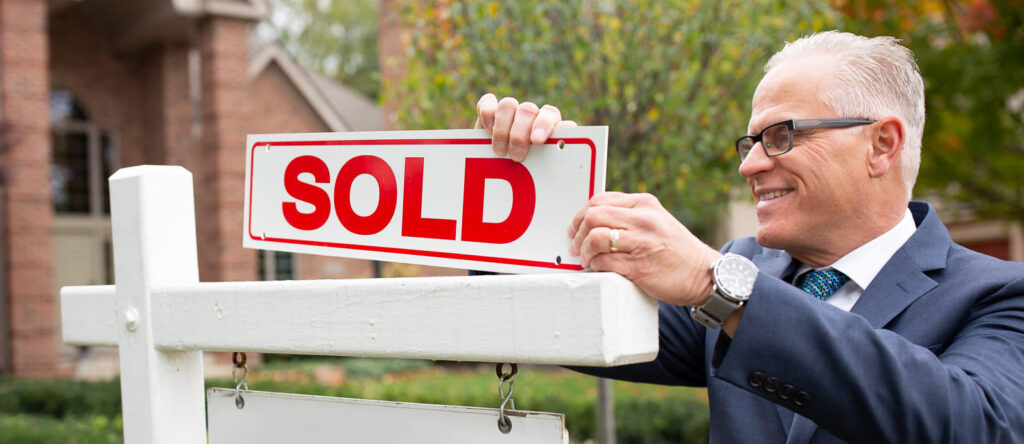 The Realtor John Goodman of John Goodman Real Estate standing behind a recently sold sign on the yard of a two story home.