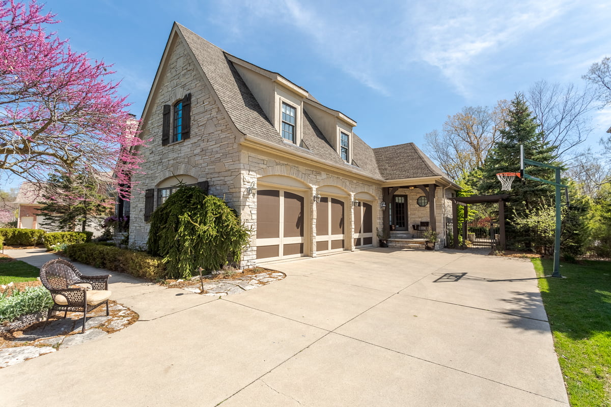Front view of a stone-clad estate home in Farmington, featuring a three-car garage, a basketball hoop on the driveway, and blooming trees surrounding the landscaped entry path.