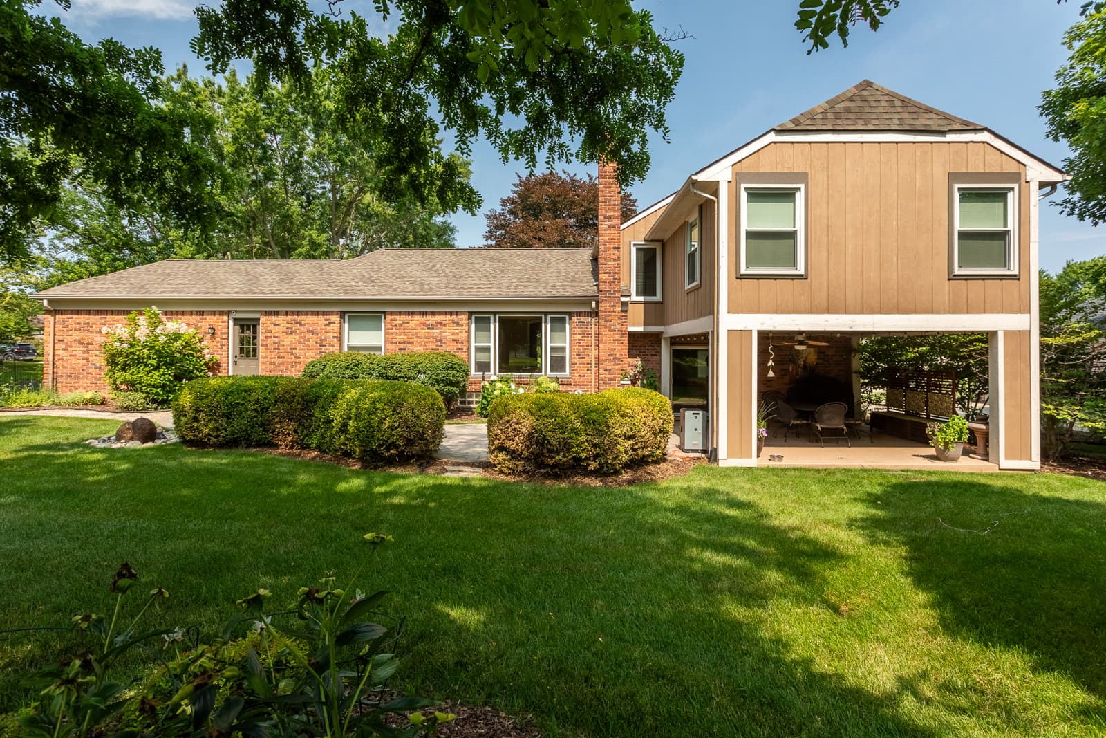 Backyard view of a Farmington, Michigan home with manicured landscaping, red brick exterior, and covered patio lounge beneath an extended upper level.